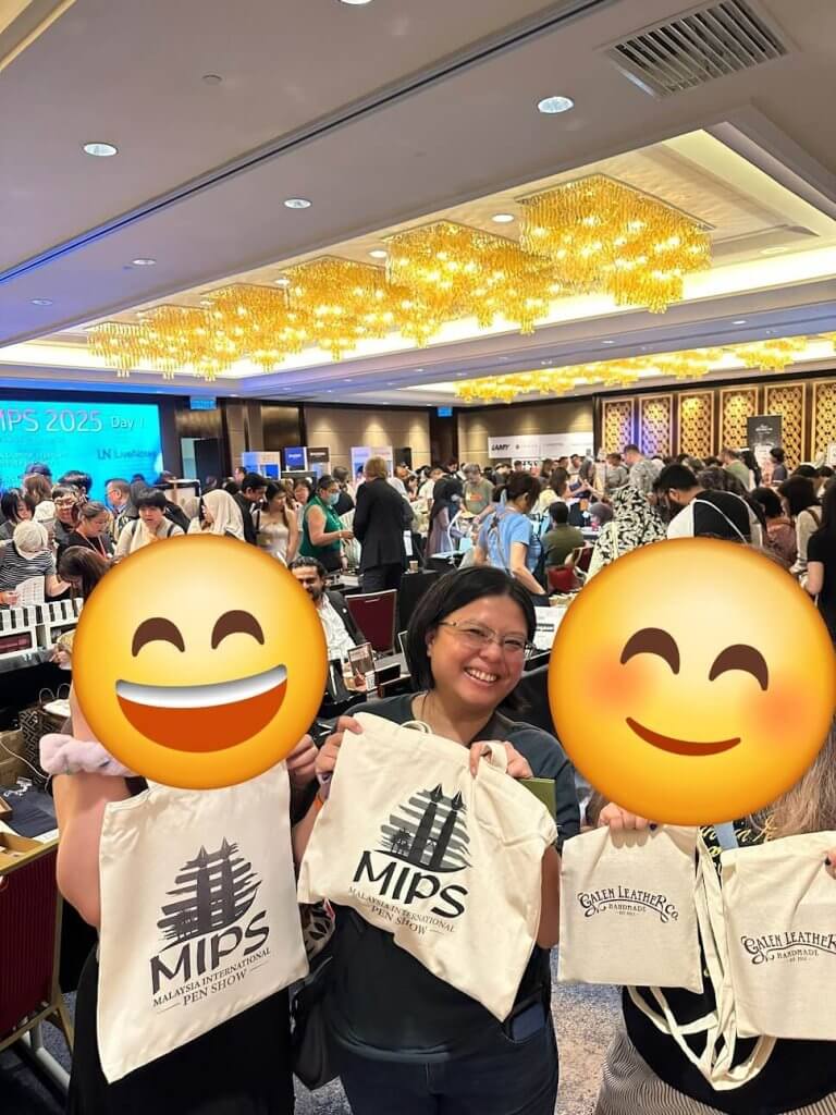 The author holds up her Malaysia International Pen Show bag with her colleagues. Their faces have been covered by a sticker